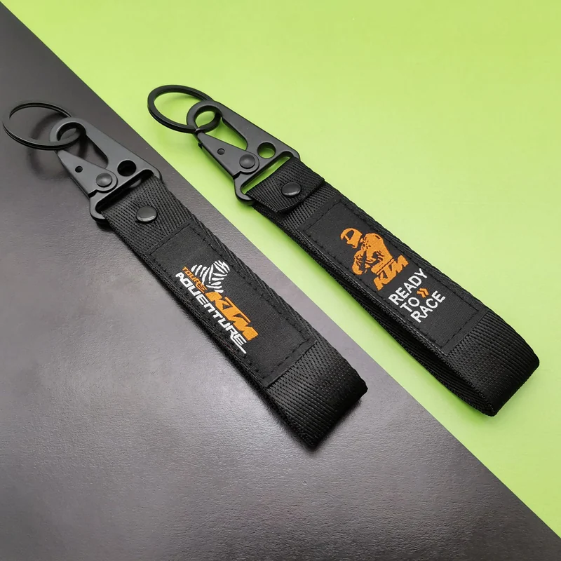 Two black short motorcycle keychain lanyards with orange sewn logo labels and black metal hooks on a green background