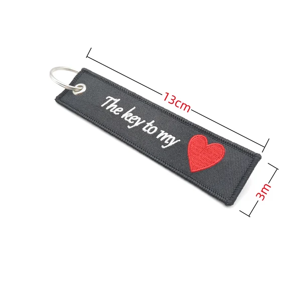 Black fabric key tag with white text and red embroidered heart showing 13 cm length and 3 cm width