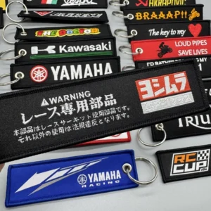 Black embroidered key tag with white warning text and red graphic displayed over assorted racing keychains