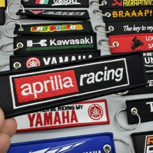 Hand holding a red and black embroidered racing key tag above assorted motorcycle keychains