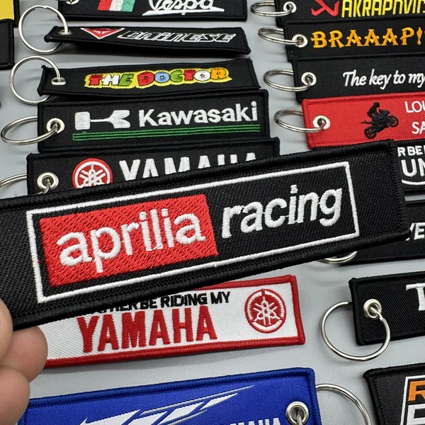 Hand holding a red and black embroidered racing key tag above assorted motorcycle keychains